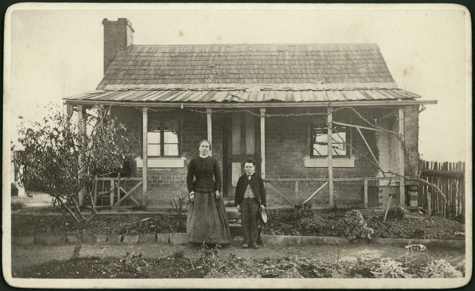 Sepia tone photograph of woman and boy standing in front of a single-storey brick cottage