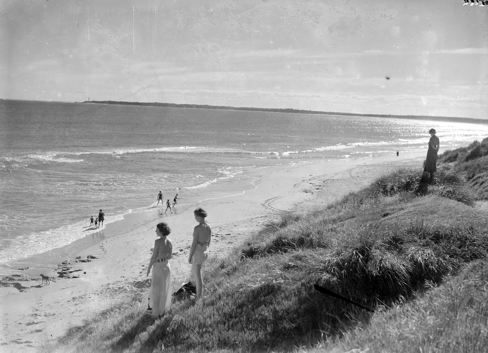 Black and white photograph of three women on a cliff overlooking beach, several people below playing at water's edge