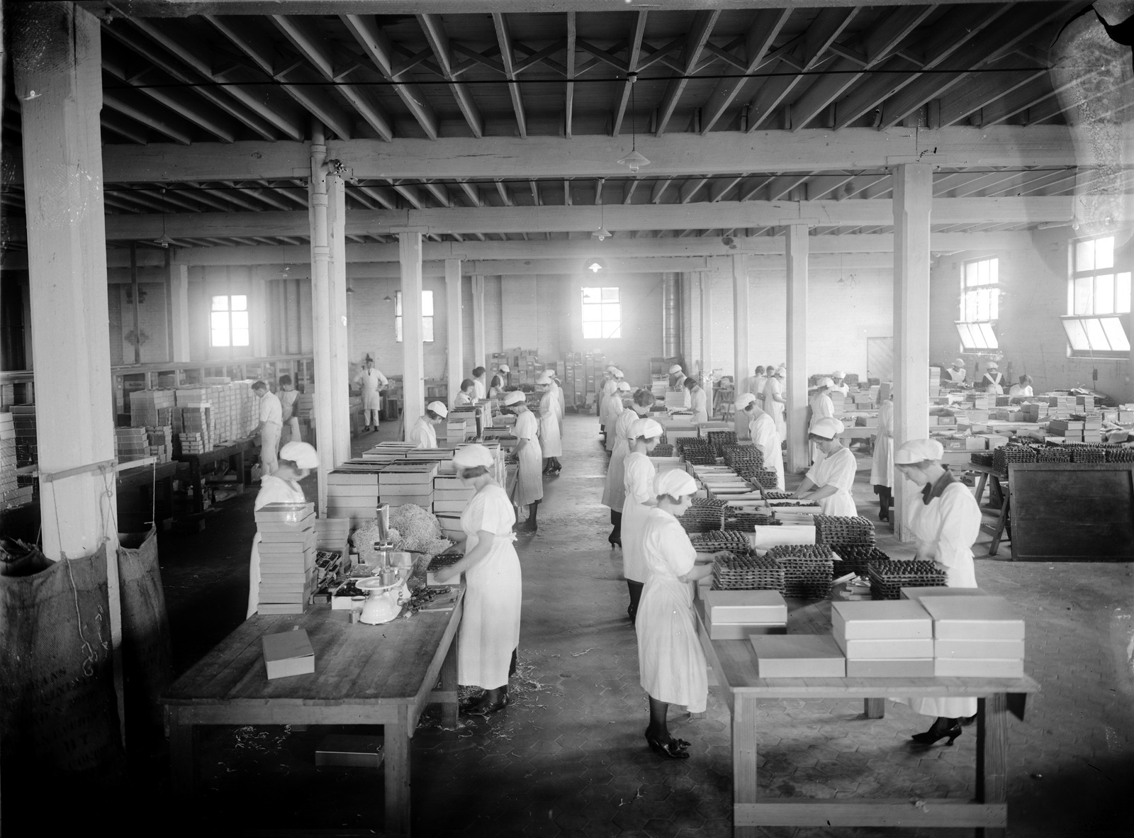 Black and white photograph of the confectionery packaging line at MacRobertson Chocolate factory. Women in white uniforms standing at large wooden tables, packing chocolates into boxes for sale