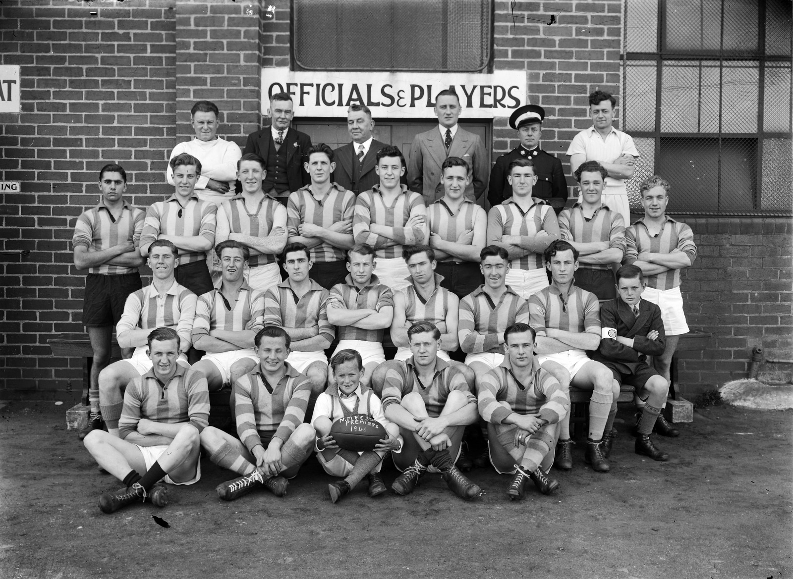 Black and white photograph of North Melbourne Football Club, 3rds. Football players assembled for a team photo in front of a brick wall, full length, front row sitting crossed legged on the ground, middle row seated, and back two rows standing