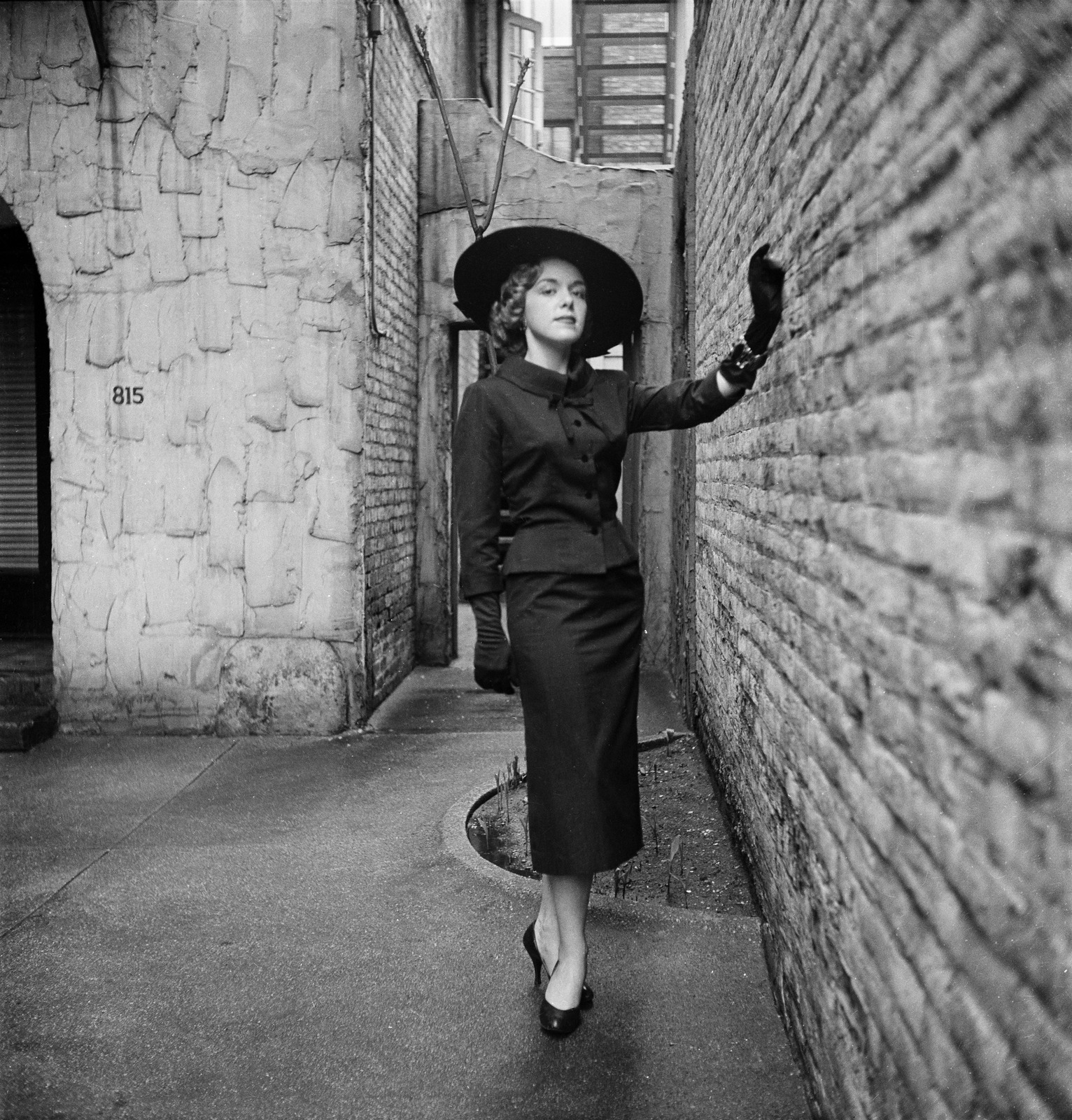 Black and white photograph of a woman wearing a long skirt, matching jacket, gloves and a hat, standing next to a brick wall