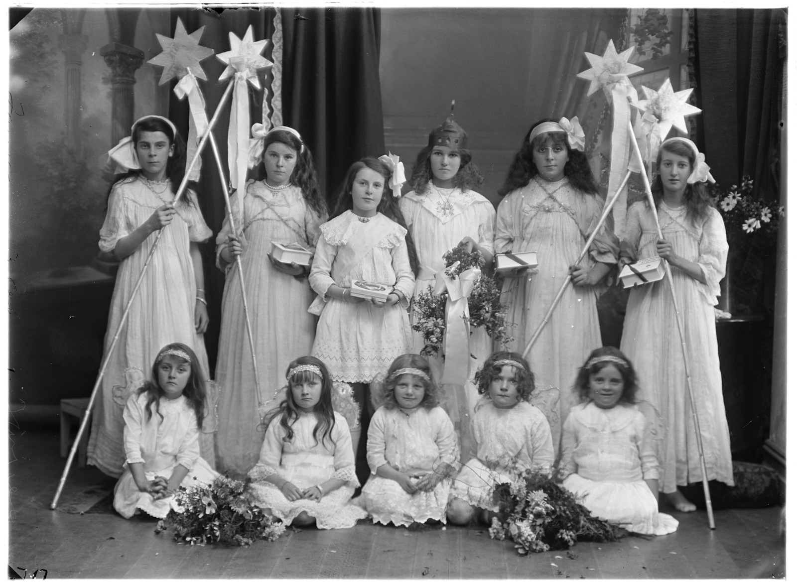 Black and white photograph showing eleven girls, whole-length, full face, younger girls seated on floor in front of standing older girls, all wearing costumes. Four girls holding staffs mounted with stars, some holding boxes of chocolates, bouquets of flowers on floor beside little girls