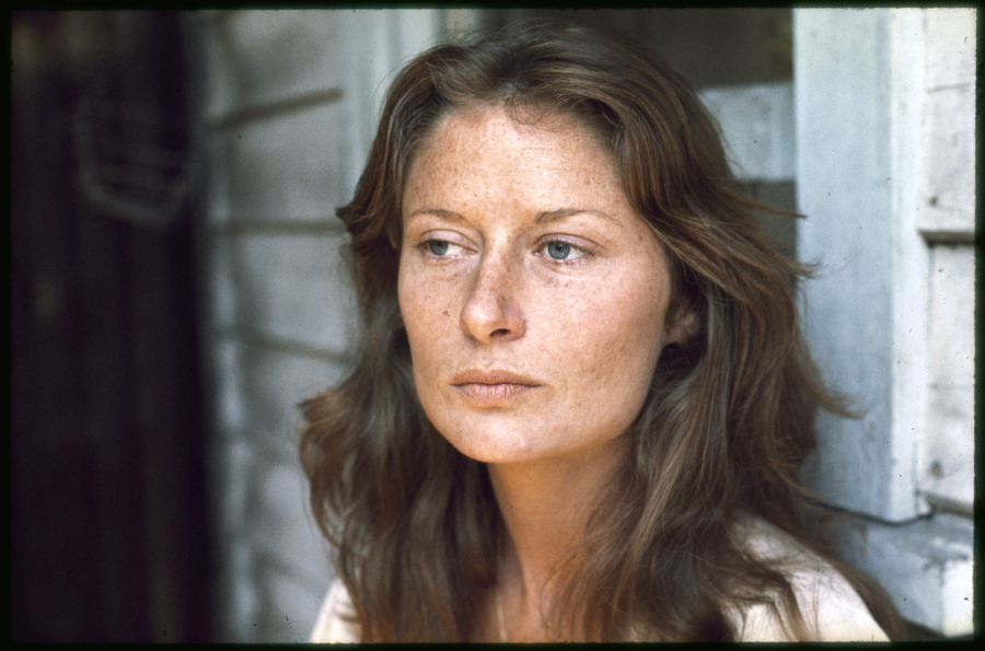 Portrait photograph of woman with long, flowing brown hair in front of a weatherboard wall and window frame