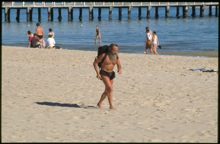 Kent Ball, former librarian at State Library Victoria, running on the beach at Albert Park in shorts, wearing a backpack. Several other people on the beach in the background and one emerging from the water, pier extending out into the water