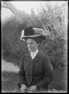 Portrait, half-length, of woman in black coat and hat seated in garden